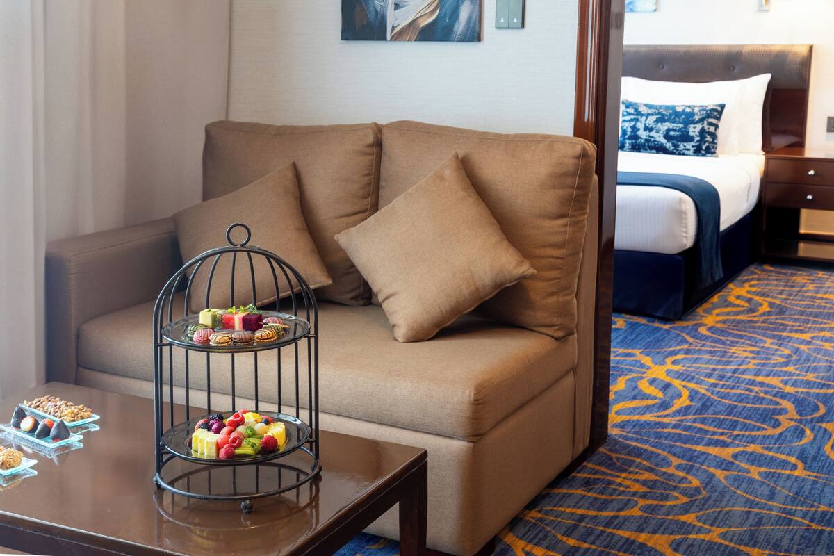 Habitación de hotel con un sofá marrón, una mesita con una bandeja de frutas y una cama con almohada azul y blanca.