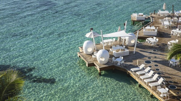 Une terrasse en bois avec des tables et des chaises blanches, un grand parasol et une eau d’un bleu limpide.