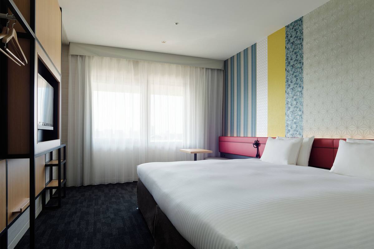 Ein Hotelzimmer mit einem großen Bett, einem Flachbildfernseher, einem Schreibtisch und einem Fenster mit durchsichtigen Vorhängen.