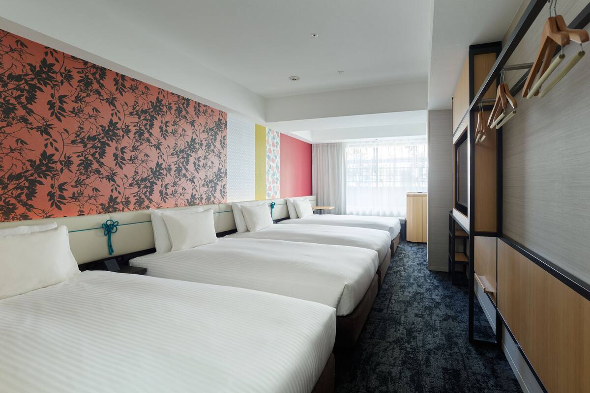 Ein Hotelzimmer mit drei Betten, einem großen Fenster und einem Kleiderschrank mit darin hängender Kleidung.