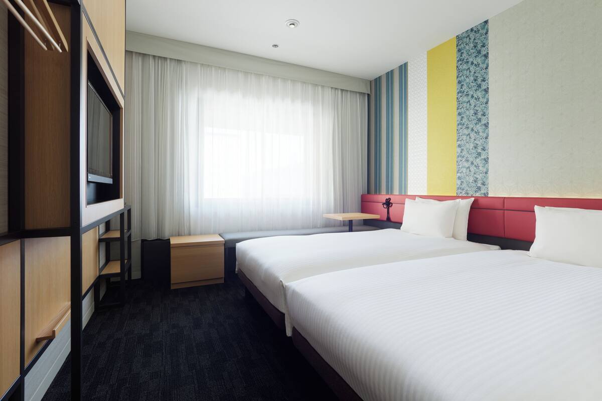 Ein Hotelzimmer mit zwei Betten, einem Schreibtisch, einem Kleiderschrank und einem großen Fenster mit Vorhängen.