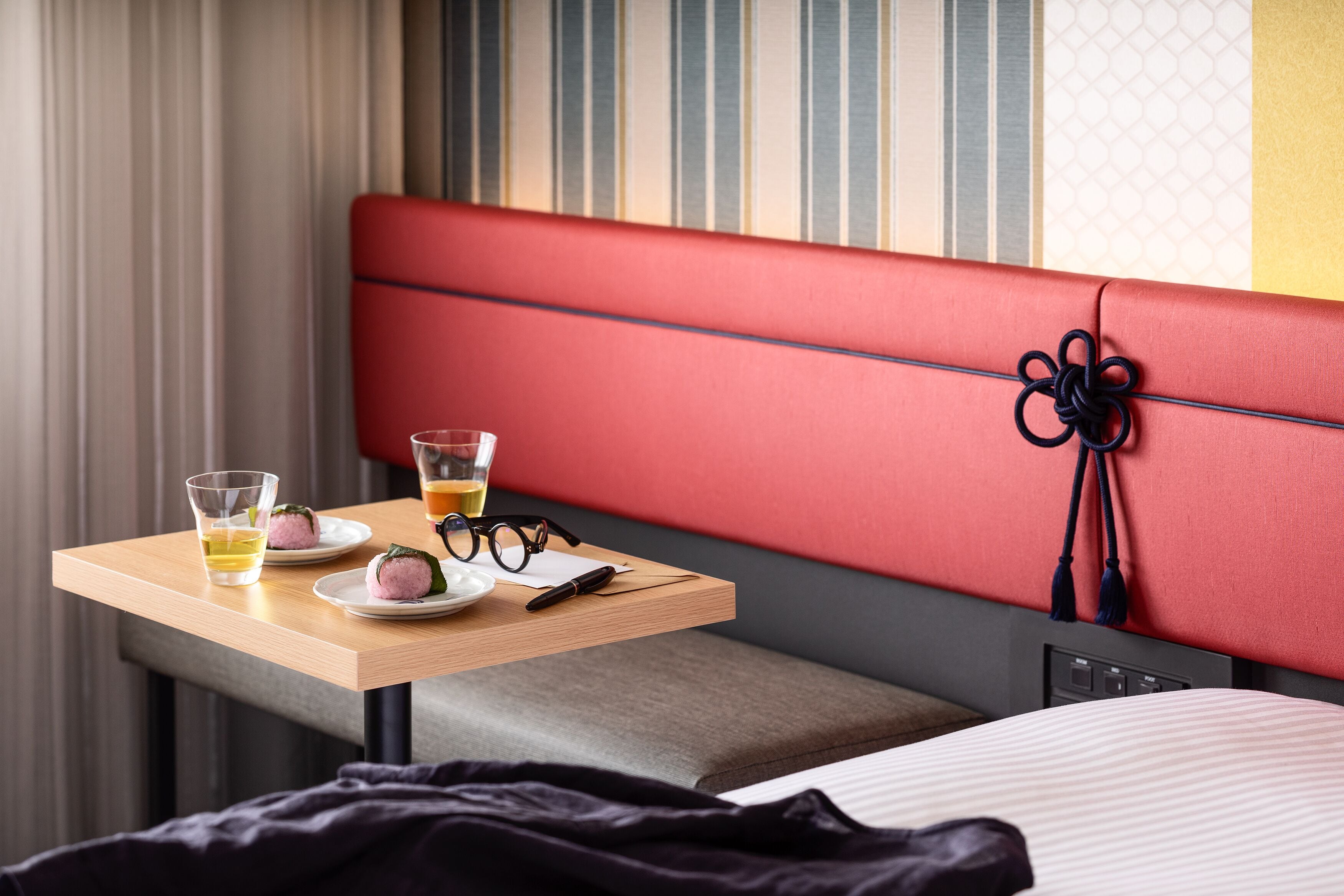 A hotel room with a red cushioned bench, a small table with glasses and a drink, and a bed with a striped bedspread.