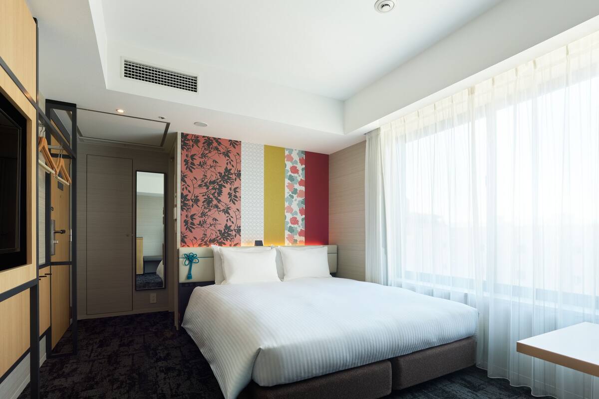 Ein modernes Hotelzimmer mit einem großen Bett, einem Schreibtisch und Blick auf die Stadt durch transparente Vorhänge.