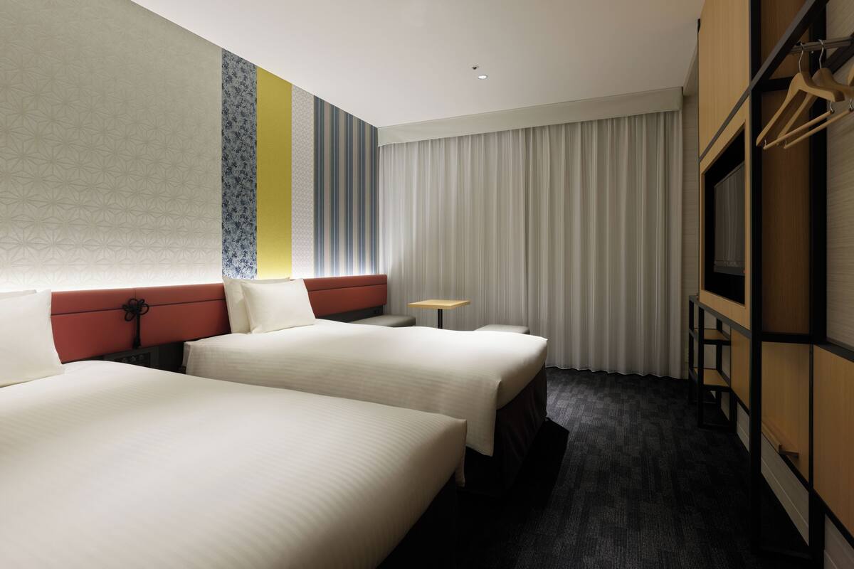 Ein Hotelzimmer mit zwei Betten, einem kleinen Tisch, einem Fernseher und einem großen Fenster mit Vorhängen.