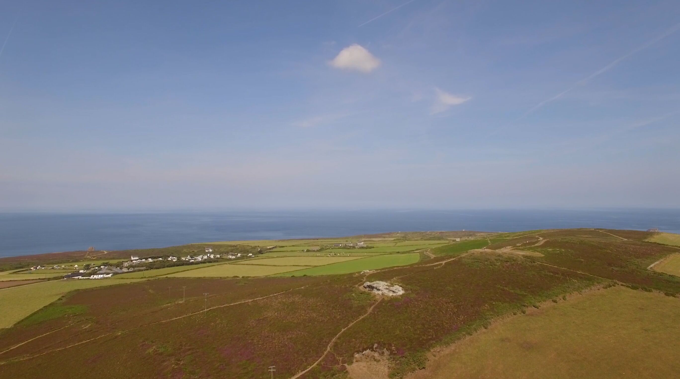 Eine Küstenlandschaft mit sanften Hügeln, einer kleinen Siedlung mit Häusern und freiem Blick auf das Meer.