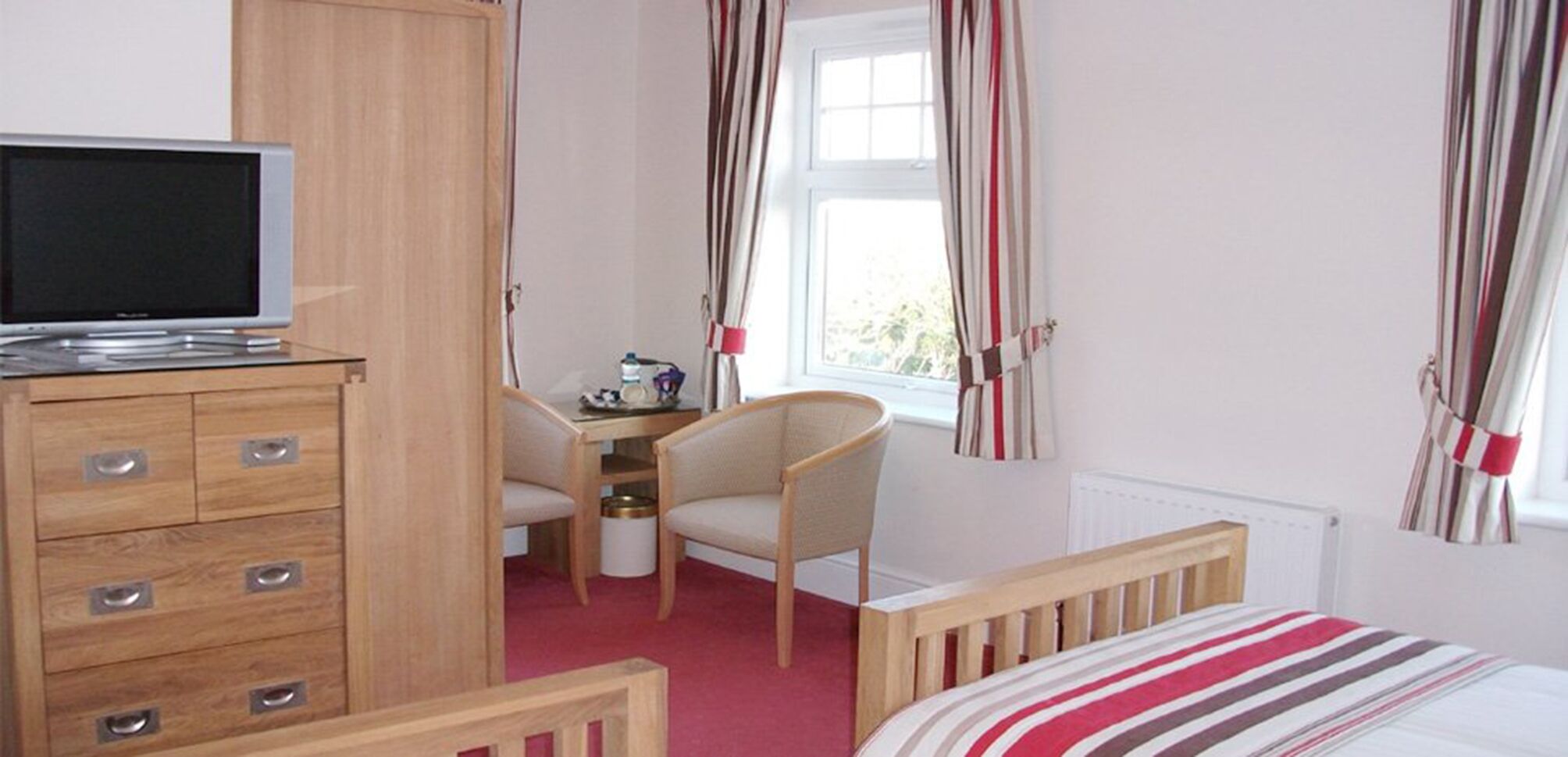A bedroom with a wooden dresser, a desk with a chair, a bed with striped bedding, and curtains on the windows.