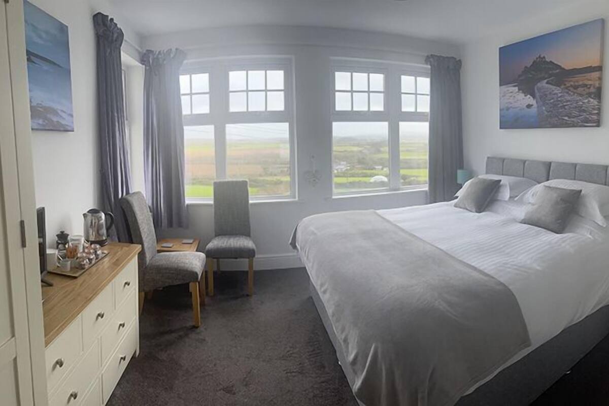 A bedroom with a large bed, a desk with a chair, a dresser, and a view of the countryside through the windows.