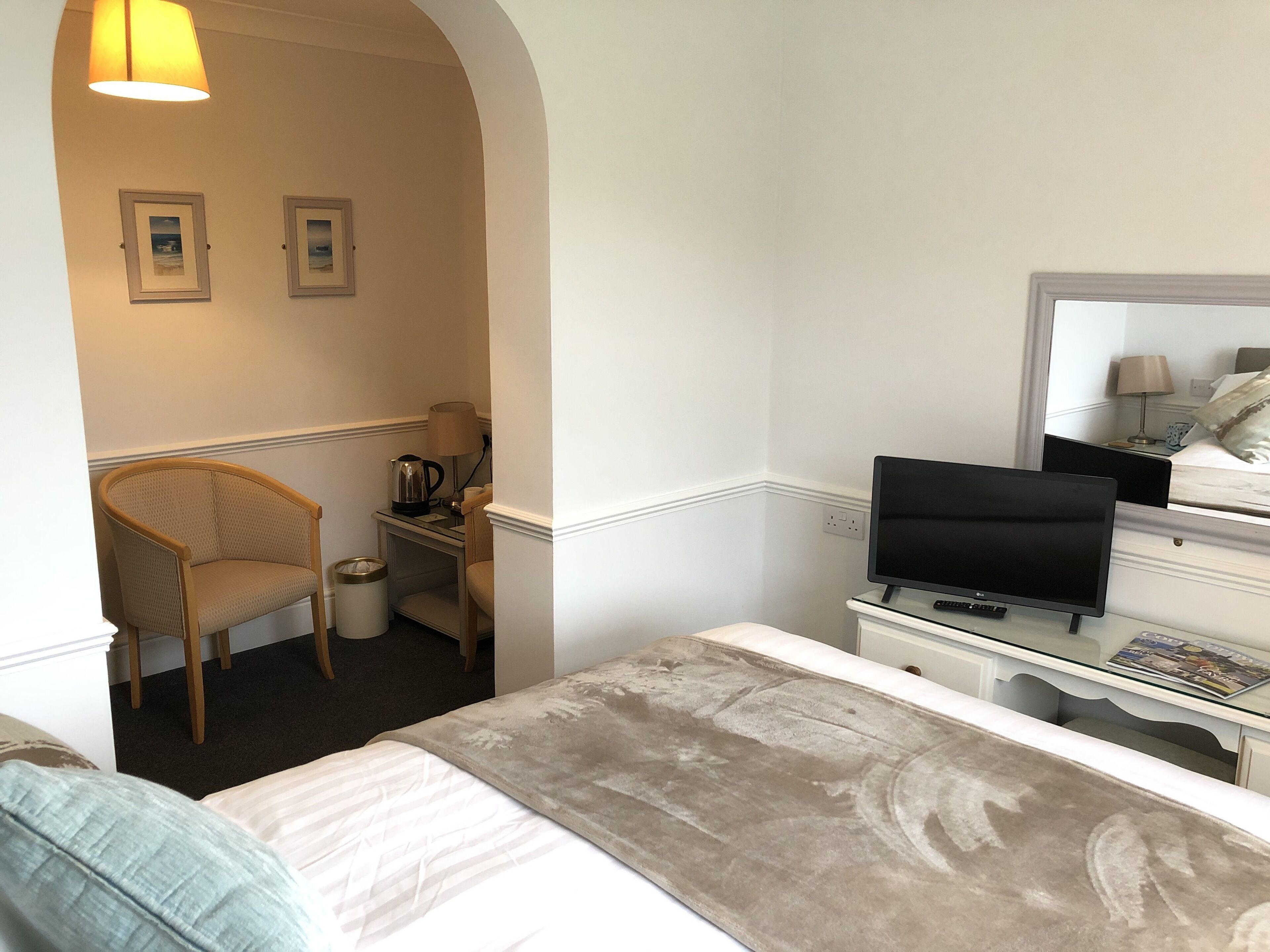 Una habitación de hotel con cama, una silla, escritorio con lámpara, televisor y espejo.