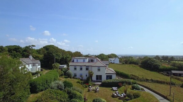 A large, white building with multiple windows and a flat roof, surrounded by lush greenery and a clear sky.