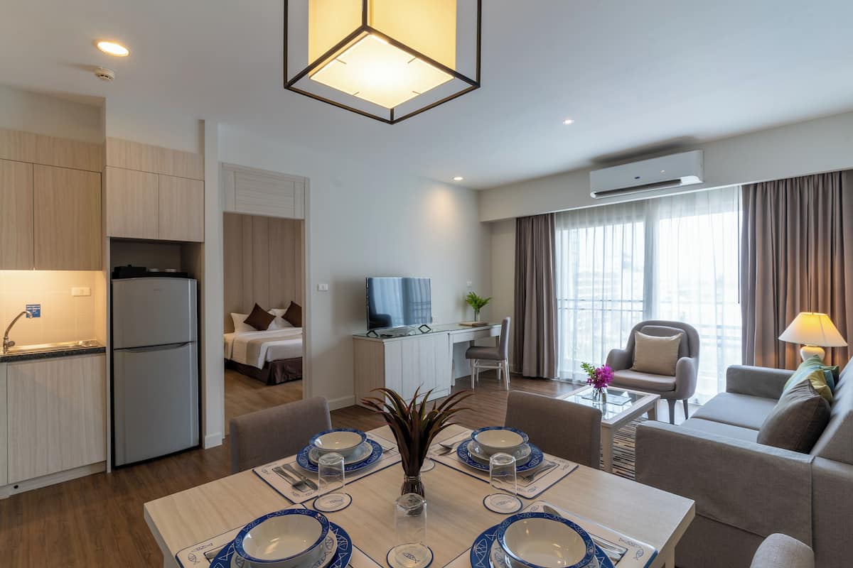 A modern hotel room with a kitchenette, a dining area with a table set for four, a living area with a sofa and armchairs, and a large window with curtains.