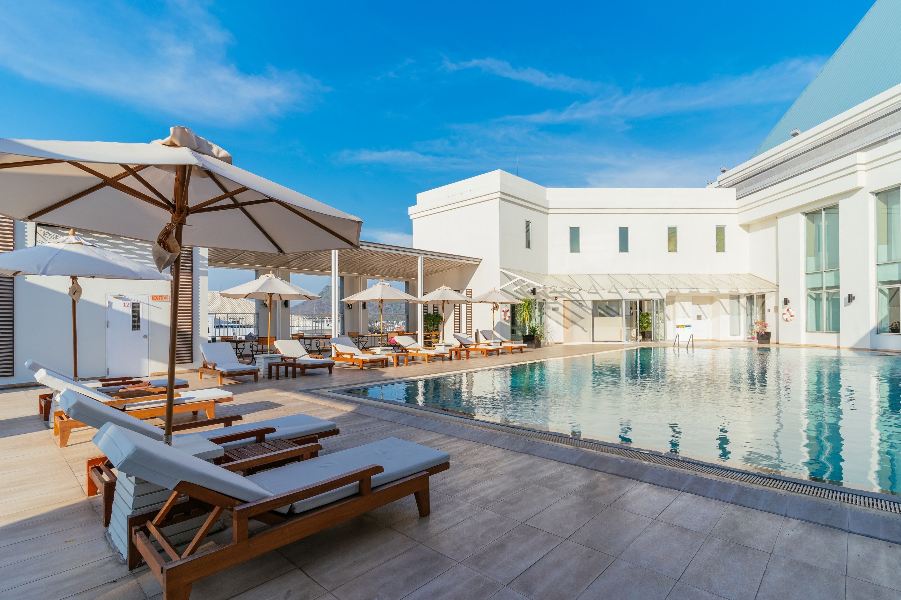 A poolside area with sun loungers and umbrellas, a clear blue sky, and a modern building in the background.