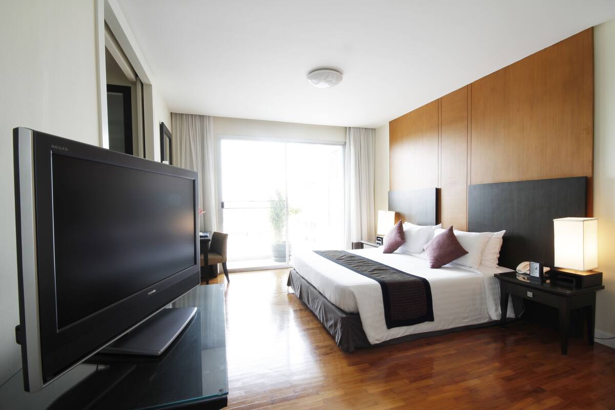 A modern hotel room with a flat-screen TV, a large bed with a wooden headboard, a bedside table with a lamp, and a view of the outdoors through a large window.
