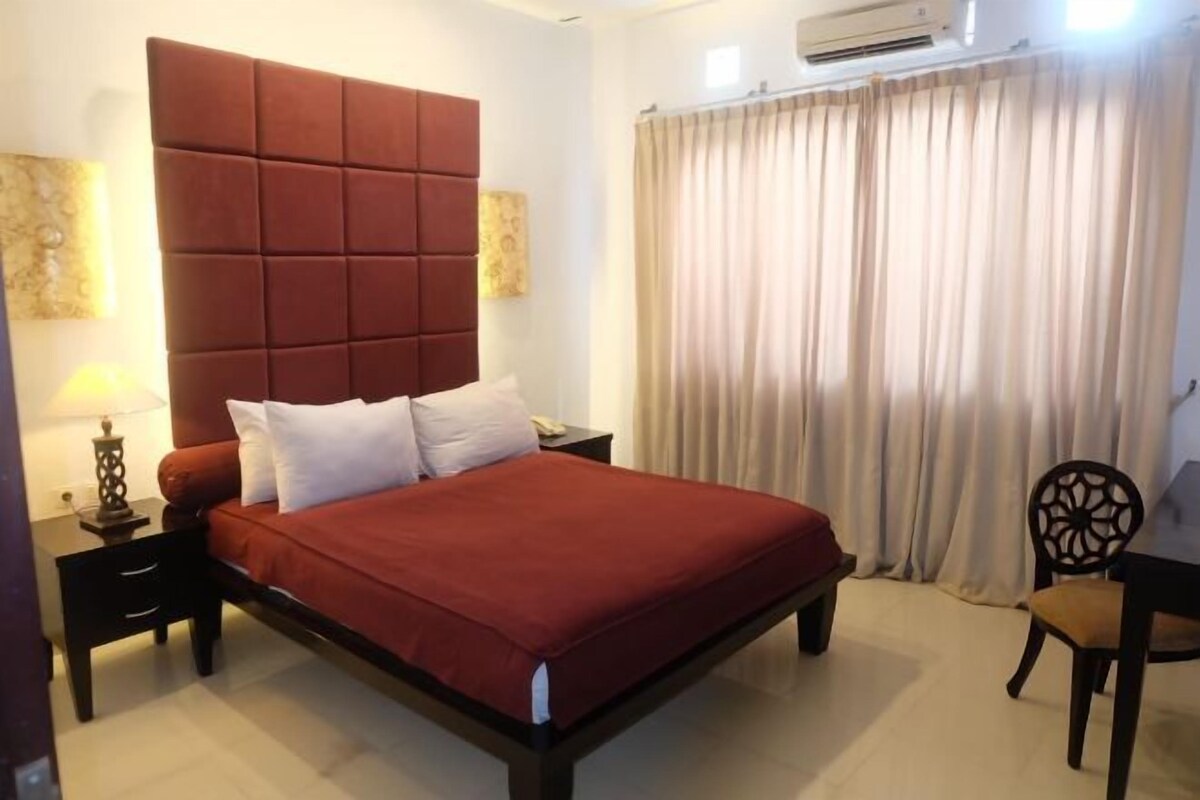 A hotel room with a large bed, a nightstand with a lamp, a chair, and a window with curtains.