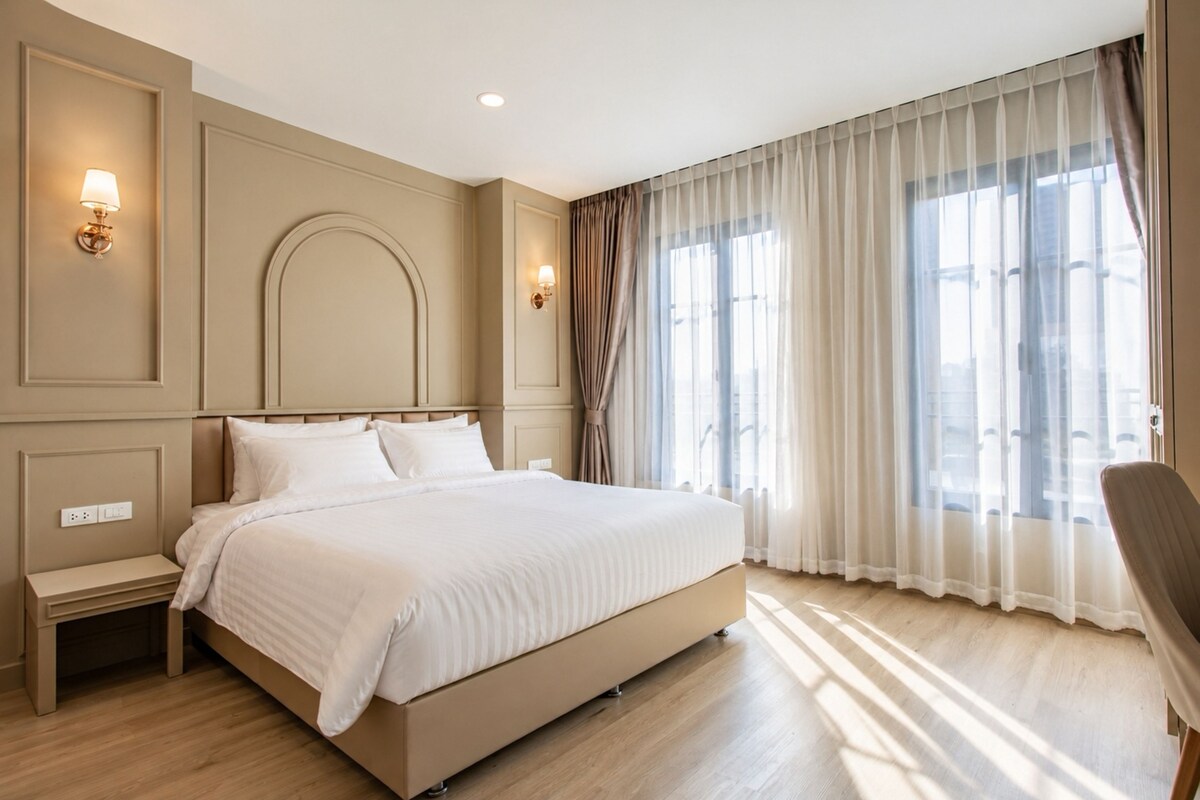 Una habitación de hotel con una cama grande, mesitas de noche, una silla y un ventanal con cortinas.