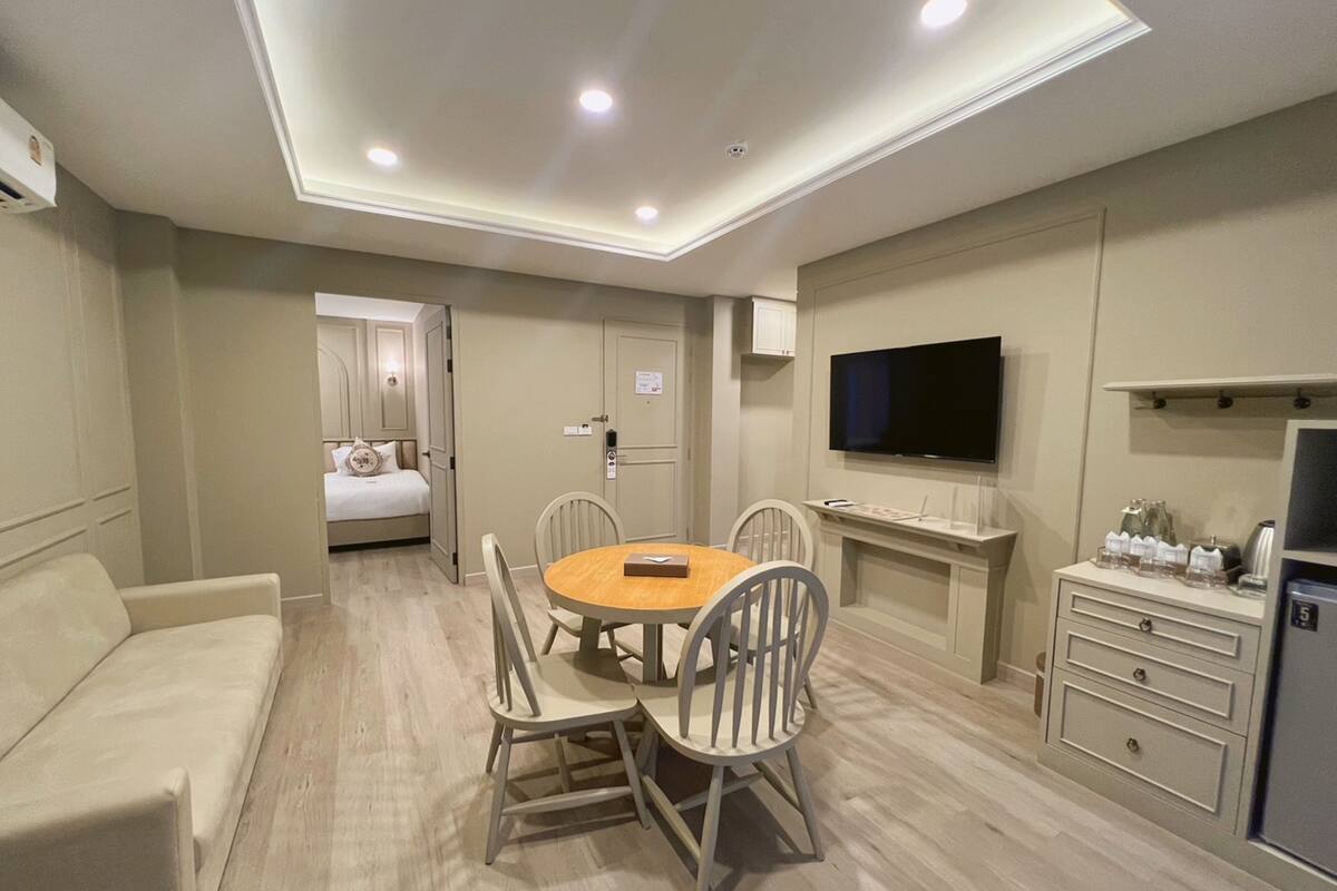 A modern hotel room with a round dining table, four chairs, a sofa, a TV, a minibar, and a door leading to a bedroom.