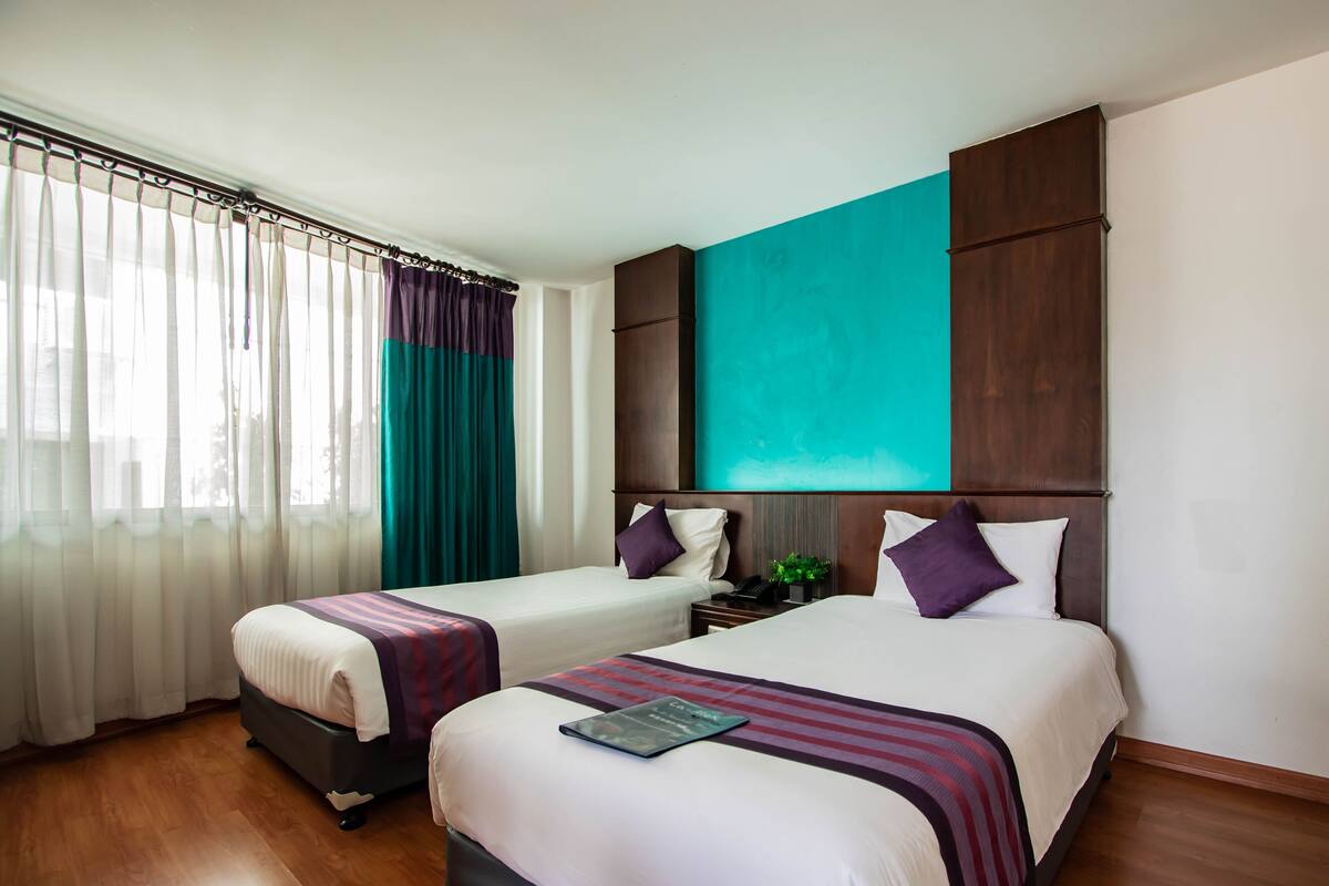 A hotel room with two beds, a large window with curtains, a wooden floor, and a large teal artwork on the wall.