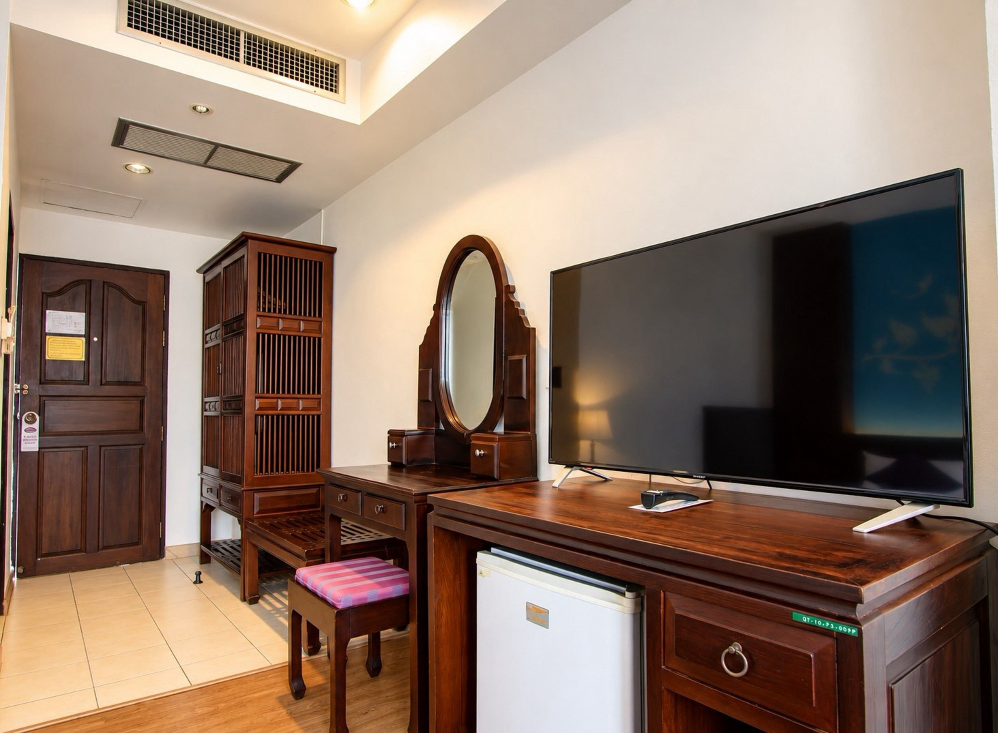 A hotel room with a wooden dresser, a television, a mirror, a small refrigerator, and a bench.