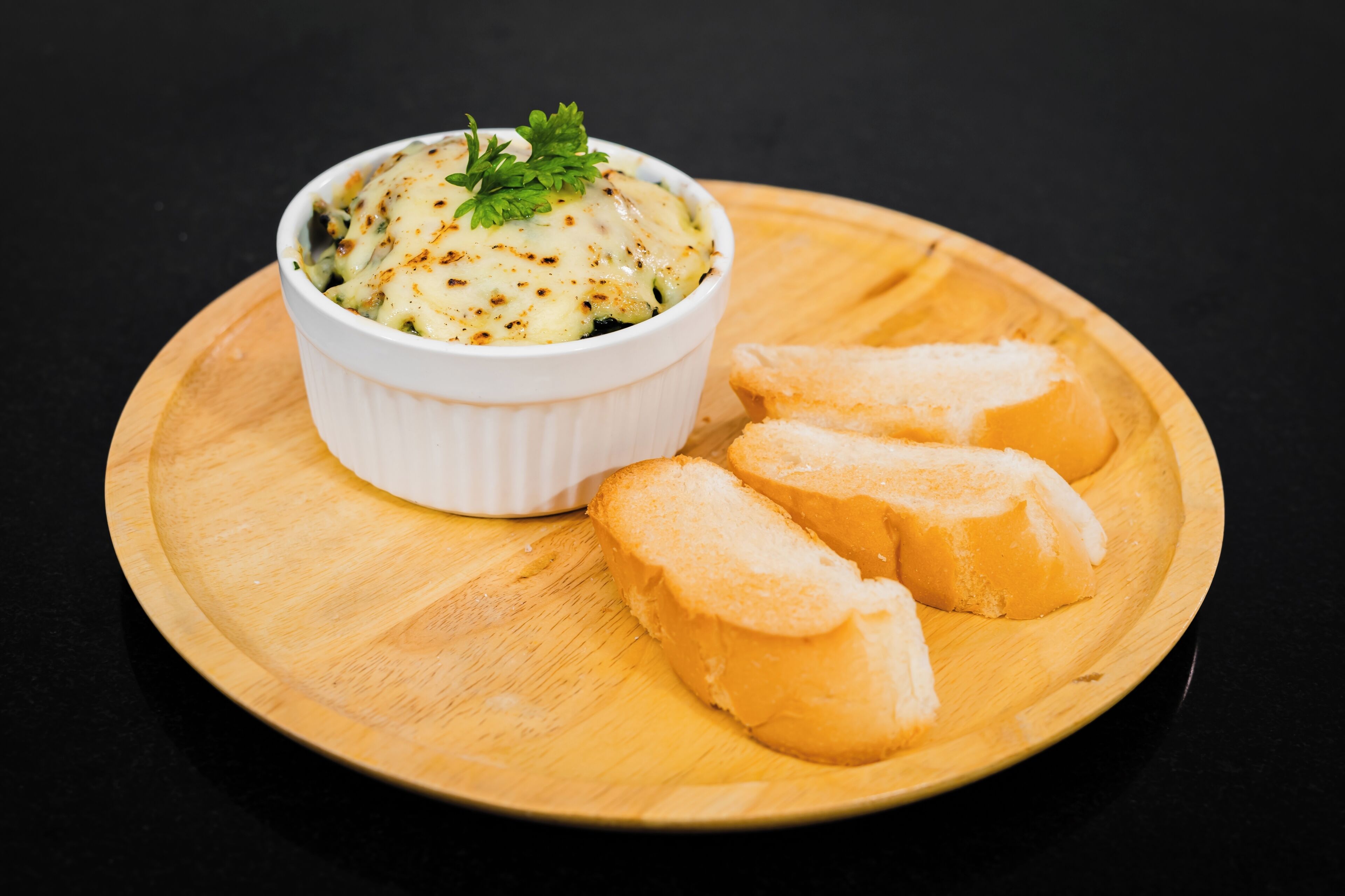 A white ceramic dish with a creamy, cheesy topping, garnished with parsley, accompanied by three slices of toasted bread on a wooden board.