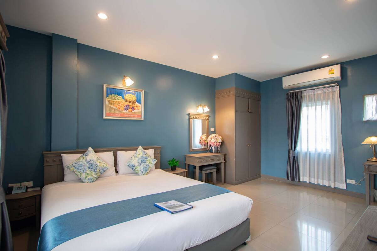 A hotel room with a large bed, a nightstand, a dresser, an air conditioning unit, and a window with curtains.