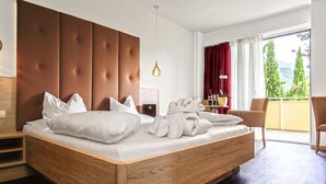 A modern hotel room with a wooden bed, a large padded headboard, a balcony with red curtains, and a view of greenery. - Anatol (Merano)