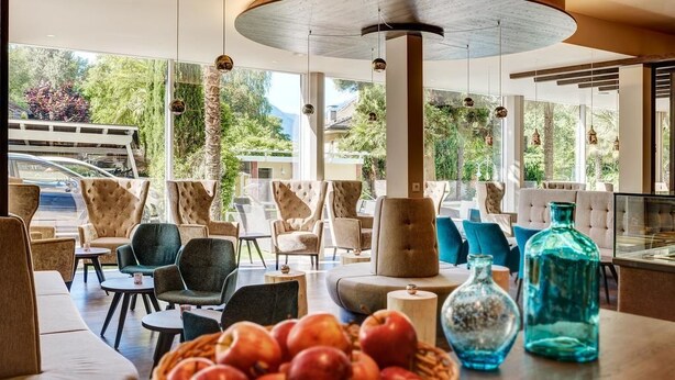 A modern lounge area with comfortable seating, a central wooden column, and a view of greenery outside. - Anatol (Merano)