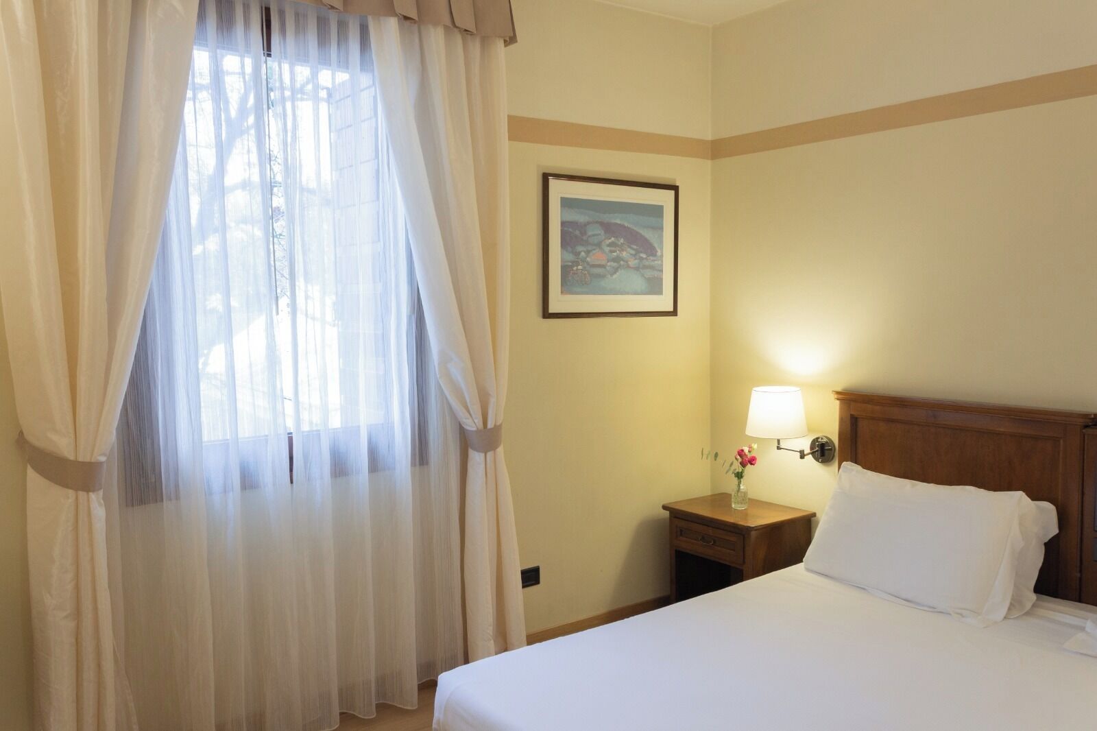 Ein Hotelzimmer mit einem Bett, einem Nachttisch mit Lampe, einem gerahmten Bild an der Wand und transparenten Vorhängen.