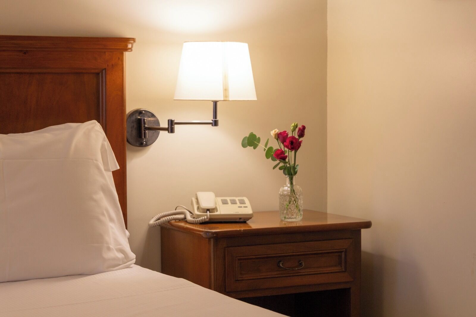 A neatly made bed with a white pillow and a wooden headboard. A bedside table with a lamp, a phone, and a vase with flowers is visible.
