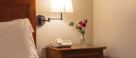 A neatly made bed with a white pillow and a wooden headboard. A bedside table with a lamp, a phone, and a vase with flowers is visible.