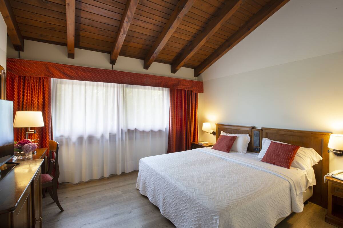 A hotel room with a wooden ceiling, a large bed with white bedding, red pillows, and two bedside lamps.