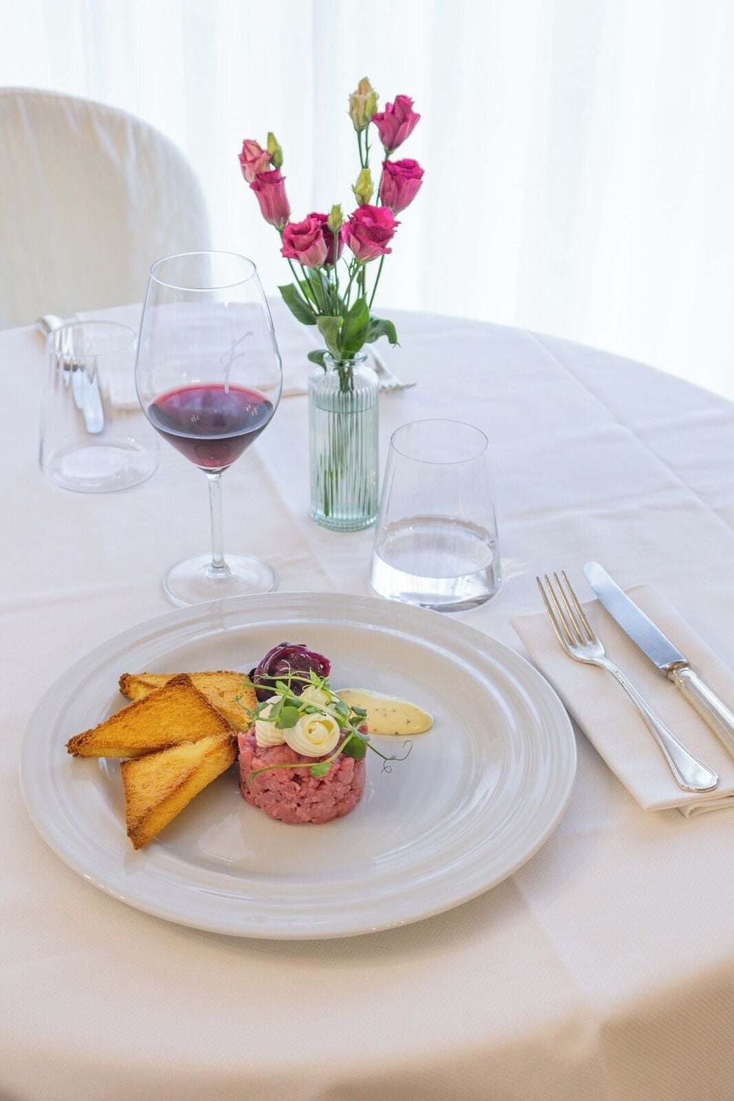 Una mesa de comedor con un plato de comida, una copa de vino tinto y un jarrón de flores rosadas.