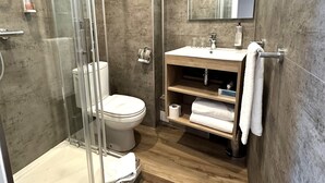 A modern bathroom with a glass-enclosed shower, a toilet, a sink with a mirror above, and wooden shelves with towels. -  Hôtel Arc en Ciel et SPA - Les Sables d'Olonne (Les Sables-d'Olonne)