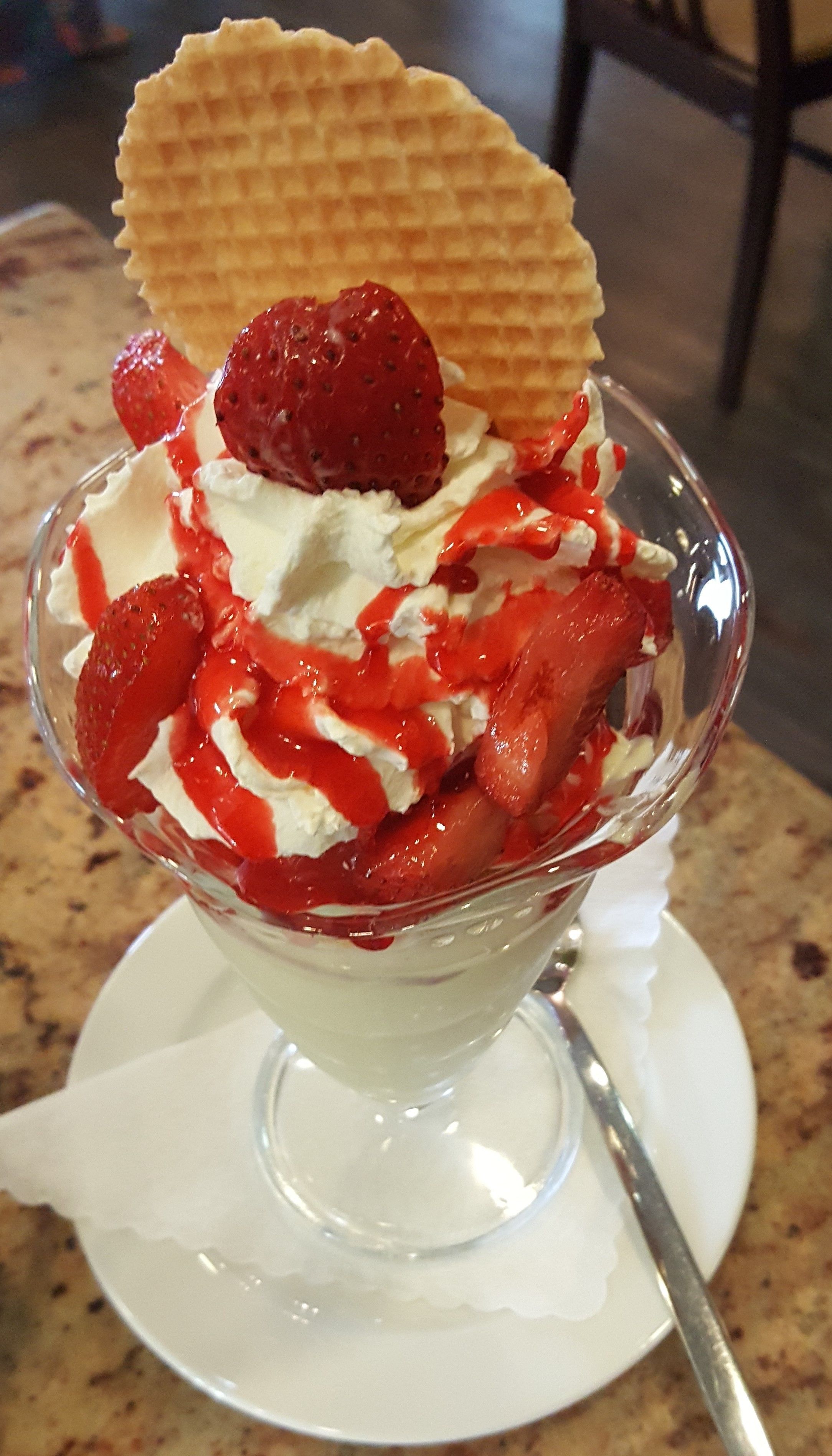 Un dessert servi dans un bol en verre transparent, garni de crème fouettée, de fraises et d’une gaufrette.