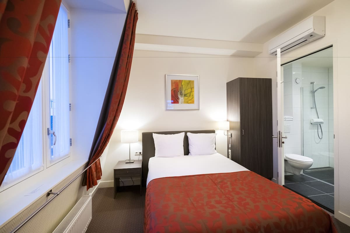 Habitación de hotel con cama, mesita de noche, ventana con cortinas, zona de ducha y un cuadro en la pared.