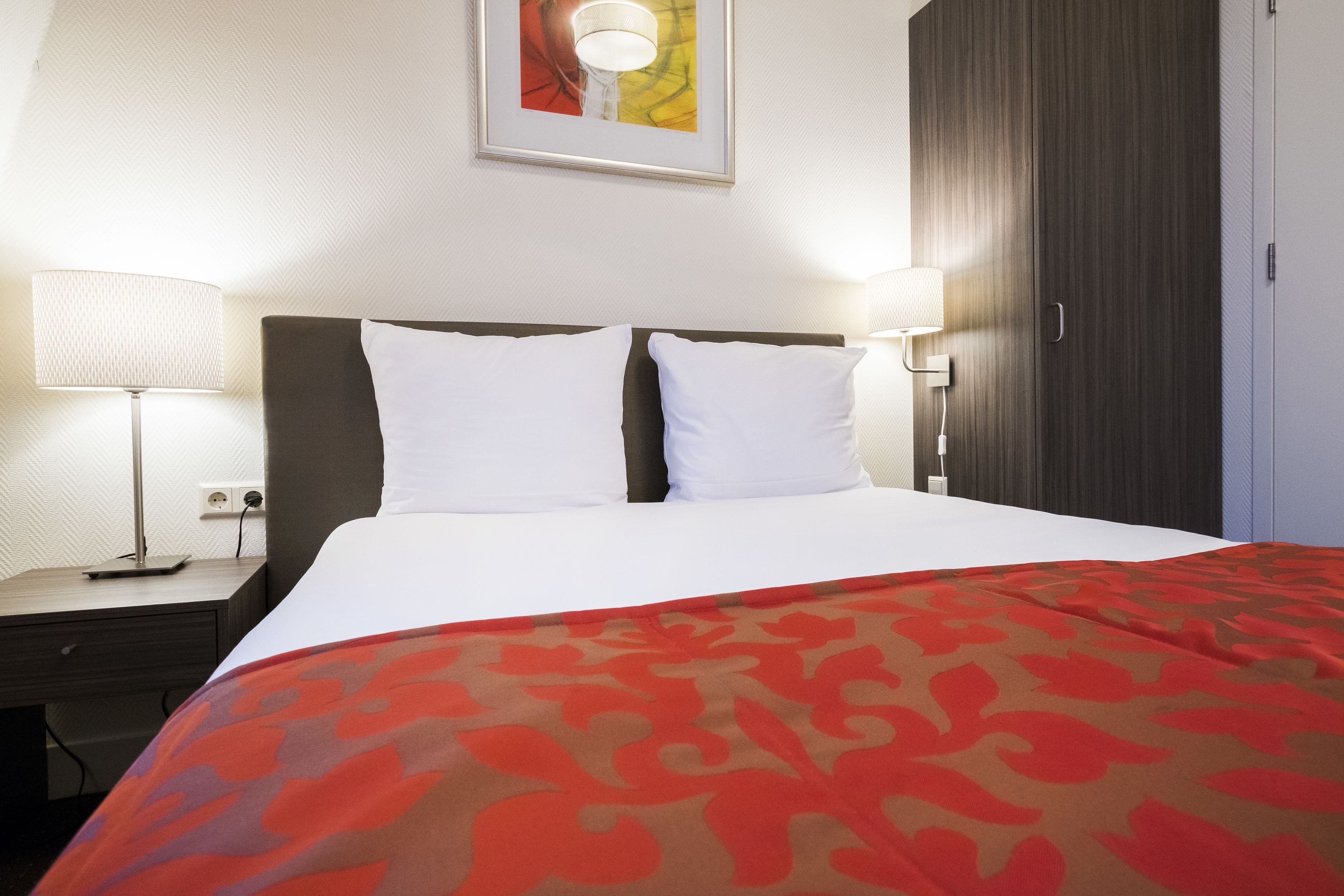 Una habitación de hotel con una cama, dos lámparas de noche, una mesita de noche, un cuadro enmarcado en la pared y un armario.