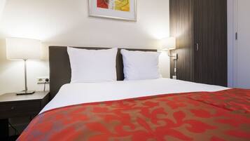 Una habitación de hotel con una cama, dos lámparas de noche, una mesita de noche, un cuadro enmarcado en la pared y un armario.