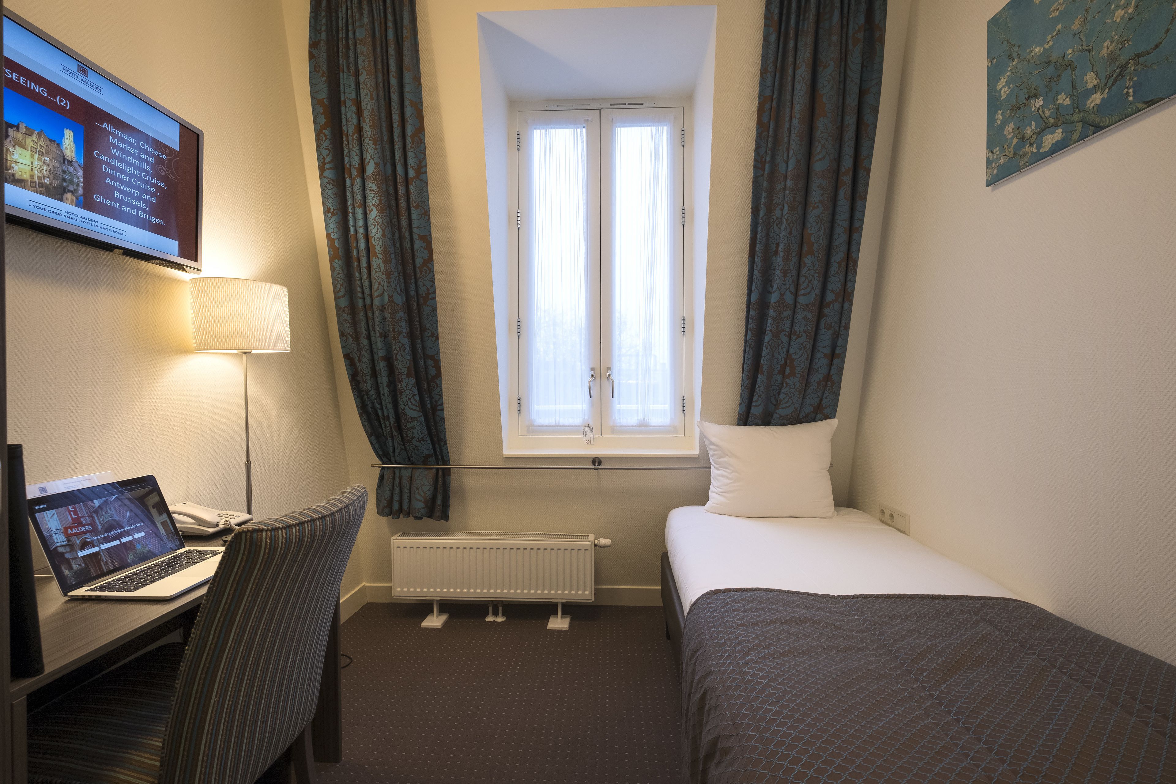 Een hotelkamer met een bed, een bureau met een laptop, een raam met gordijnen en een radiator.