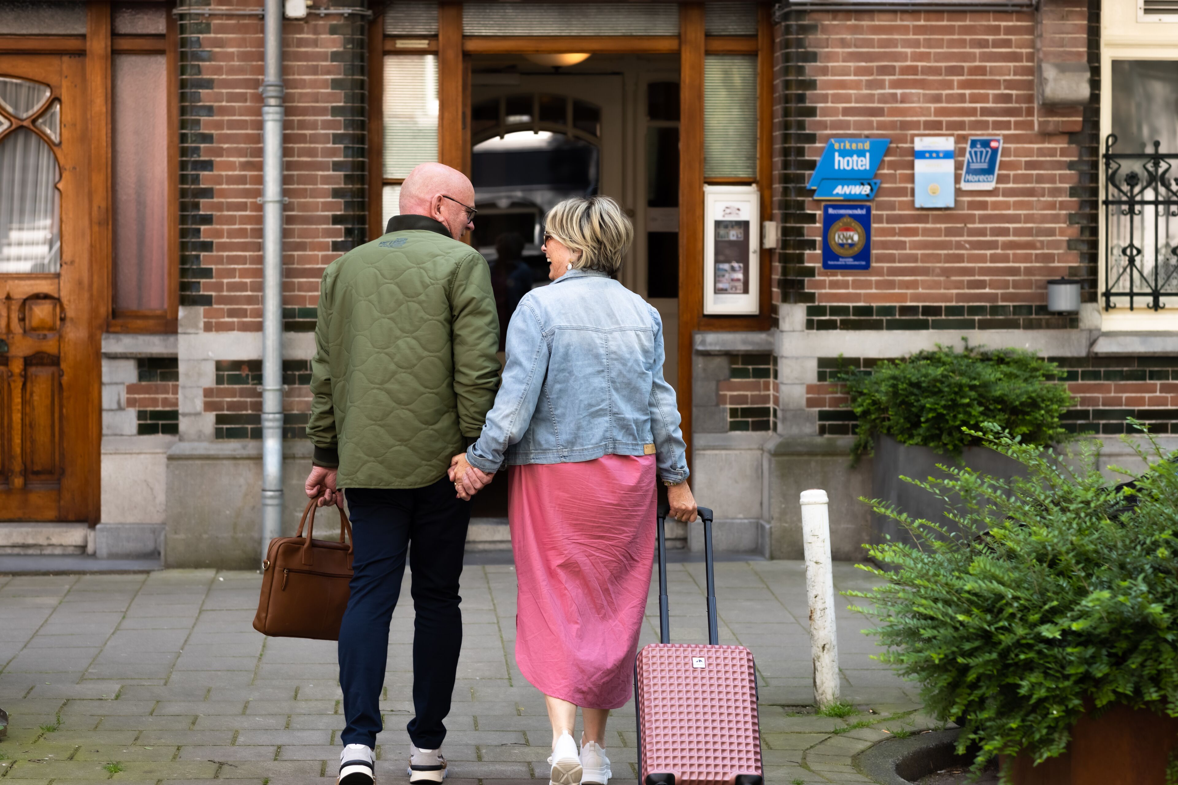 A couple walking towards a hotel entrance with a brick facade, a wooden door, and a signboard indicating the hotel name.
