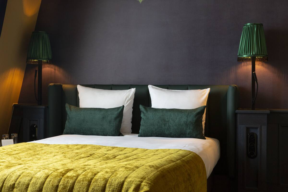 Una cama bien tendida con una colcha verde, dos almohadas verdes y dos lámparas verdes a cada lado.