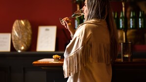 A person seated at a bar with a wine glass, a shelf of bottles, and a menu in the background. - Hotel Aalders (Amsterdam)