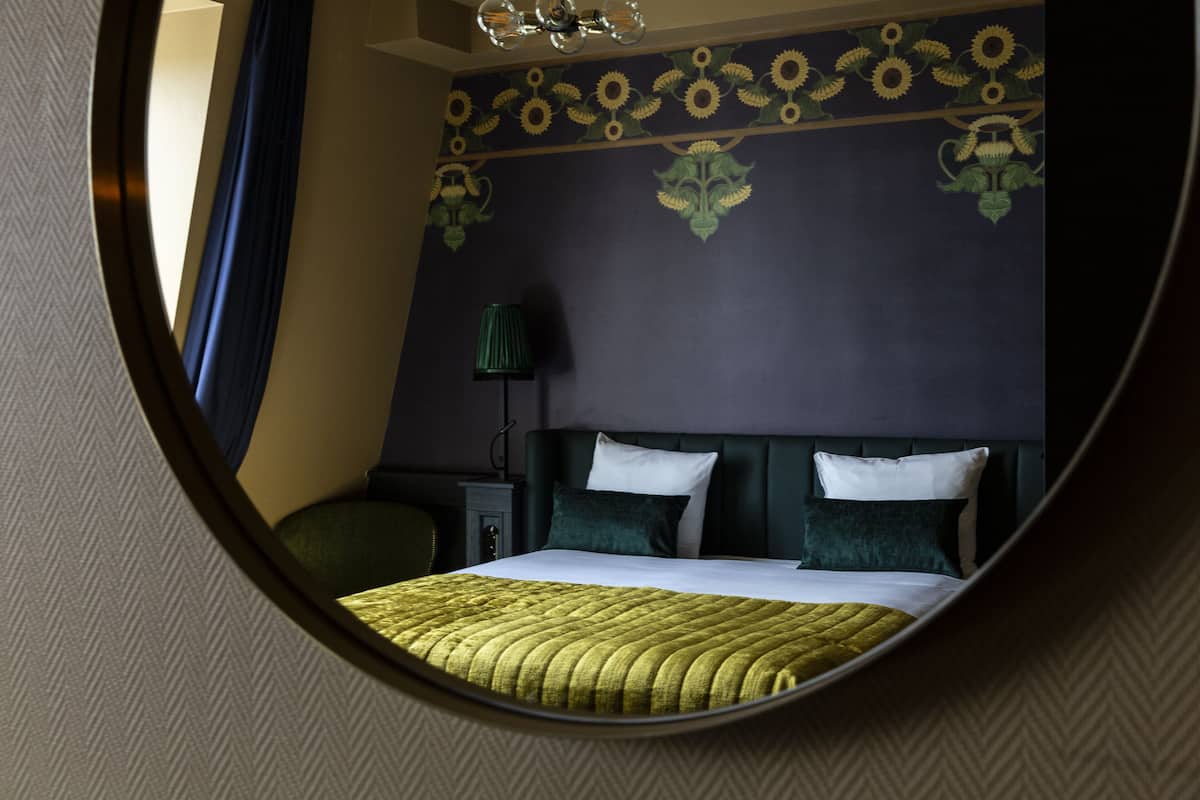 Un espejo circular que refleja un dormitorio con cama, almohadas, mesita de noche y lámpara.