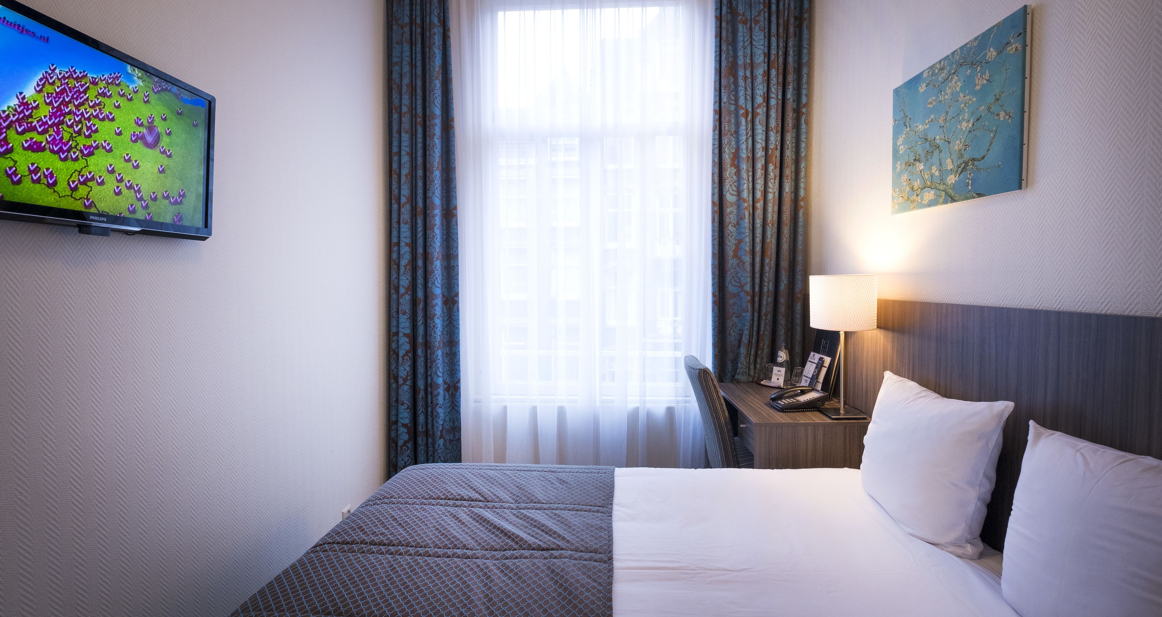 A hotel room with a bed, a desk with a lamp, a phone, and a painting on the wall.