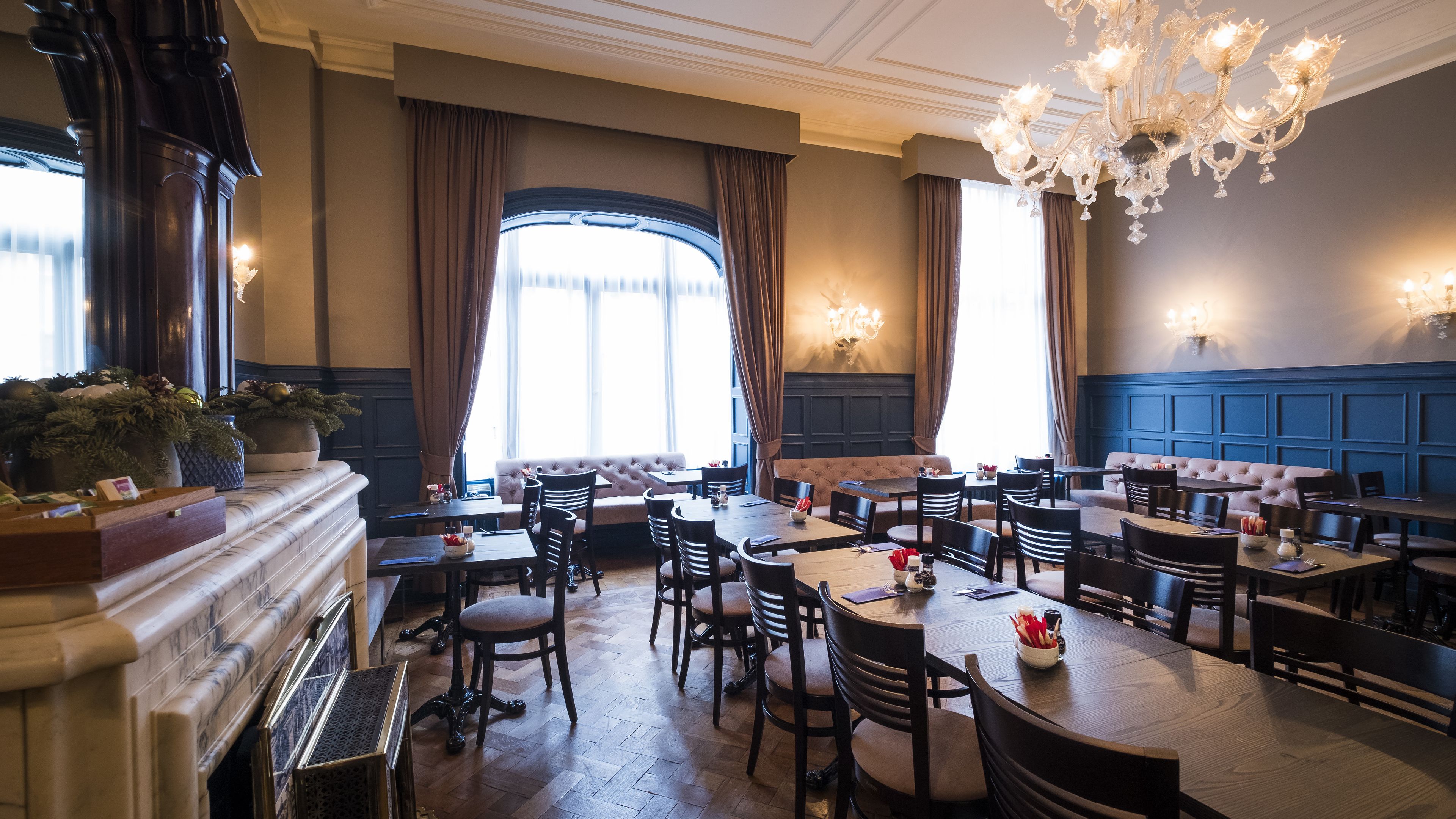 A restaurant with a classic interior, featuring wooden tables and chairs, a fireplace, and large windows with curtains.
