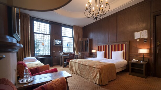 A hotel room with a large bed, two smaller beds, a desk, a chair, a table, a chandelier, and a view of the outdoors through large windows. - Hotel Aalders (Amsterdam)