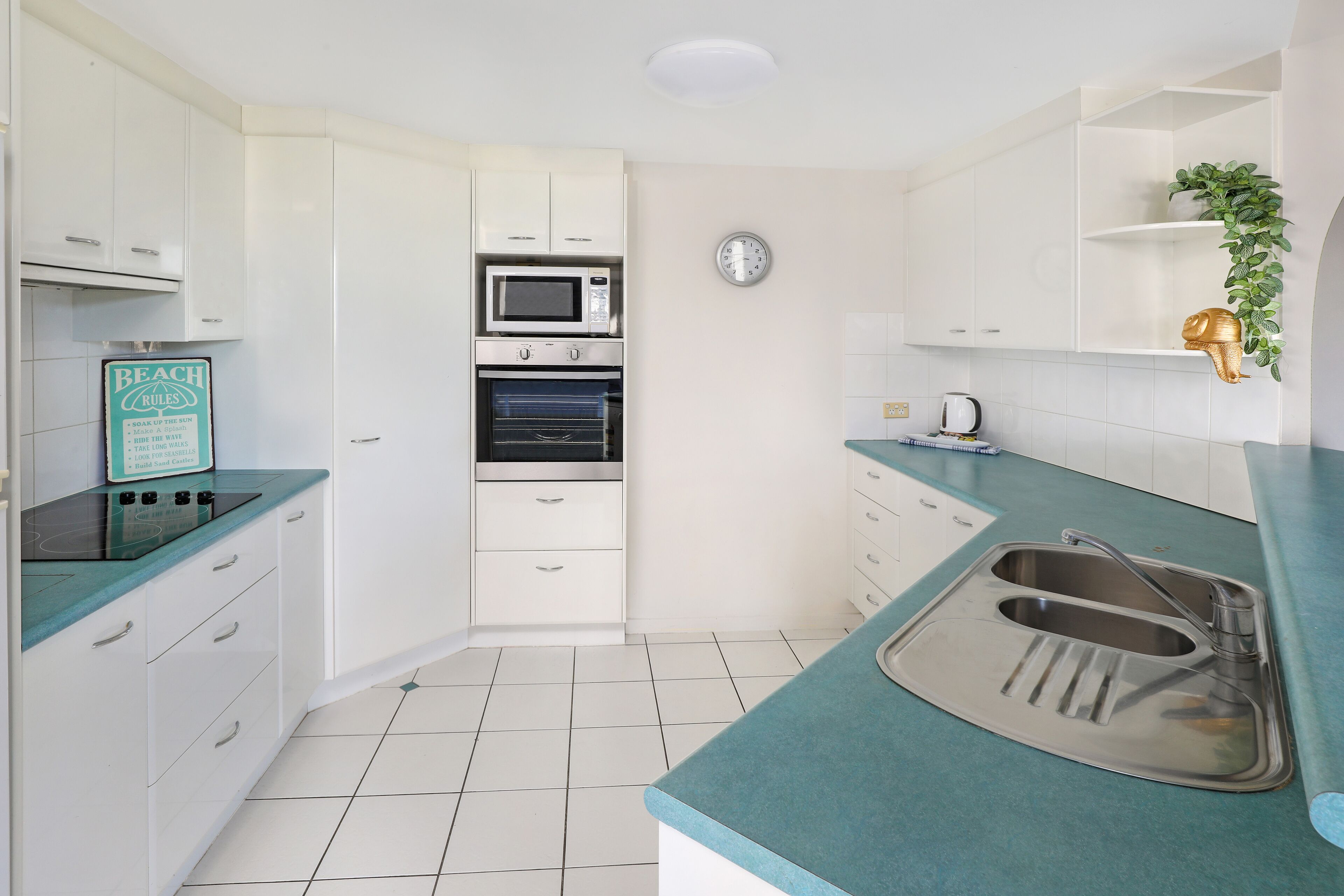 A compact kitchen with white cabinets, a microwave, an oven, a sink, and a clock on the wall.