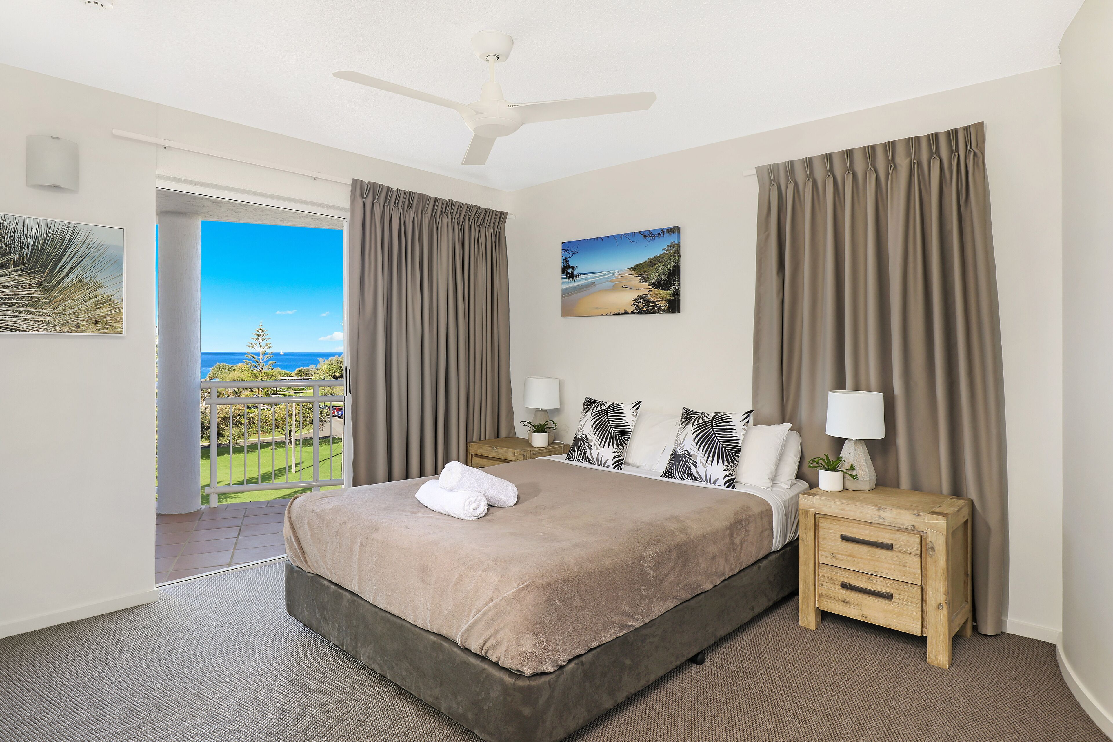 A bedroom with a bed, two bedside tables with lamps, a ceiling fan, a large window with curtains, and a view of the outdoors.