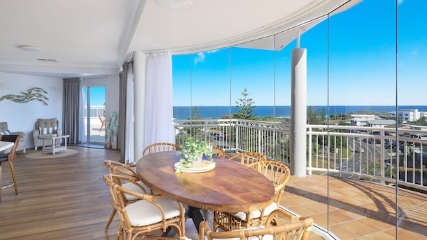 A spacious balcony with a wooden table and wicker chairs, offering a view of the ocean and cityscape.
