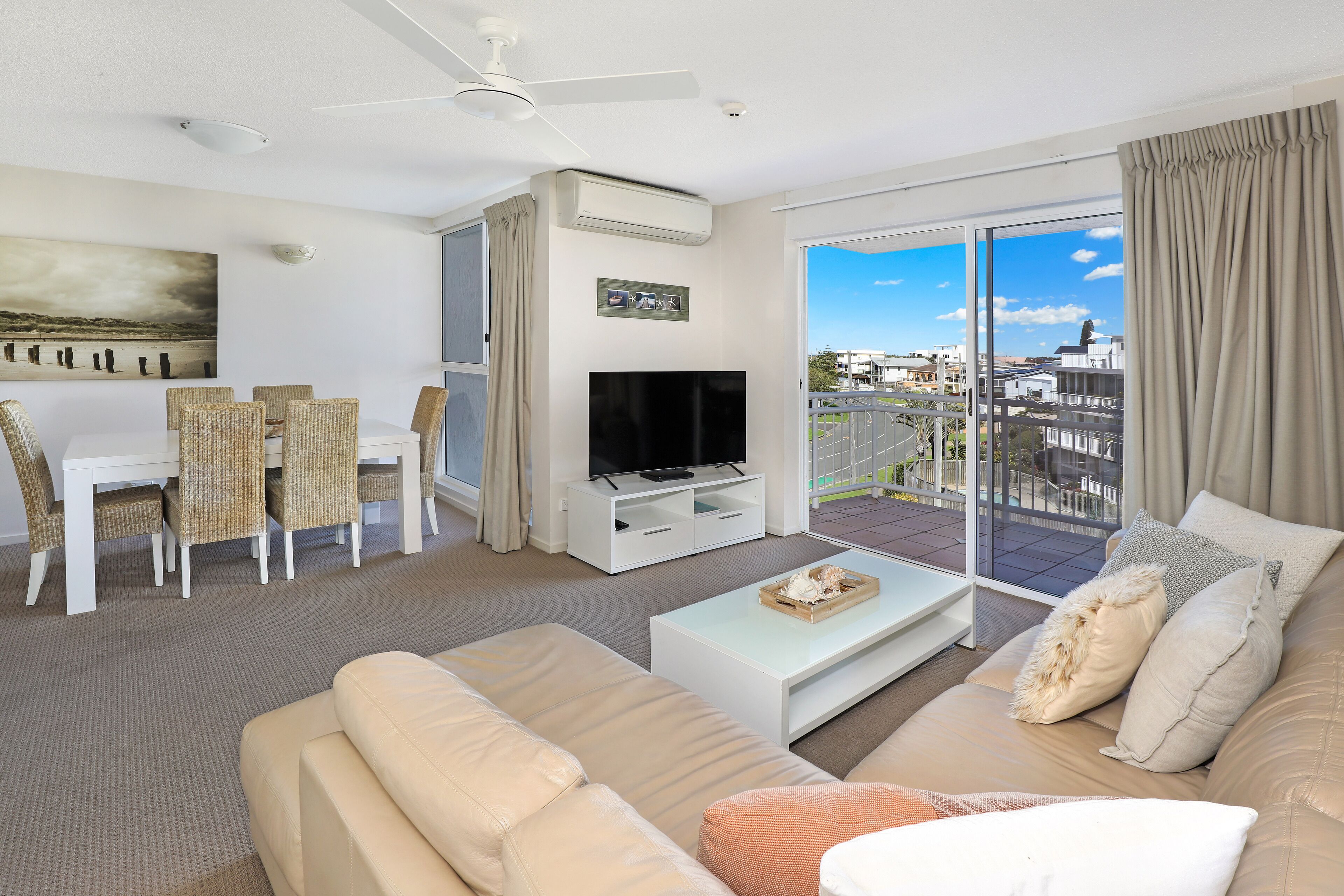 A modern living room with a flat-screen TV, a sofa, a dining table with chairs, and a balcony with a view of the city.