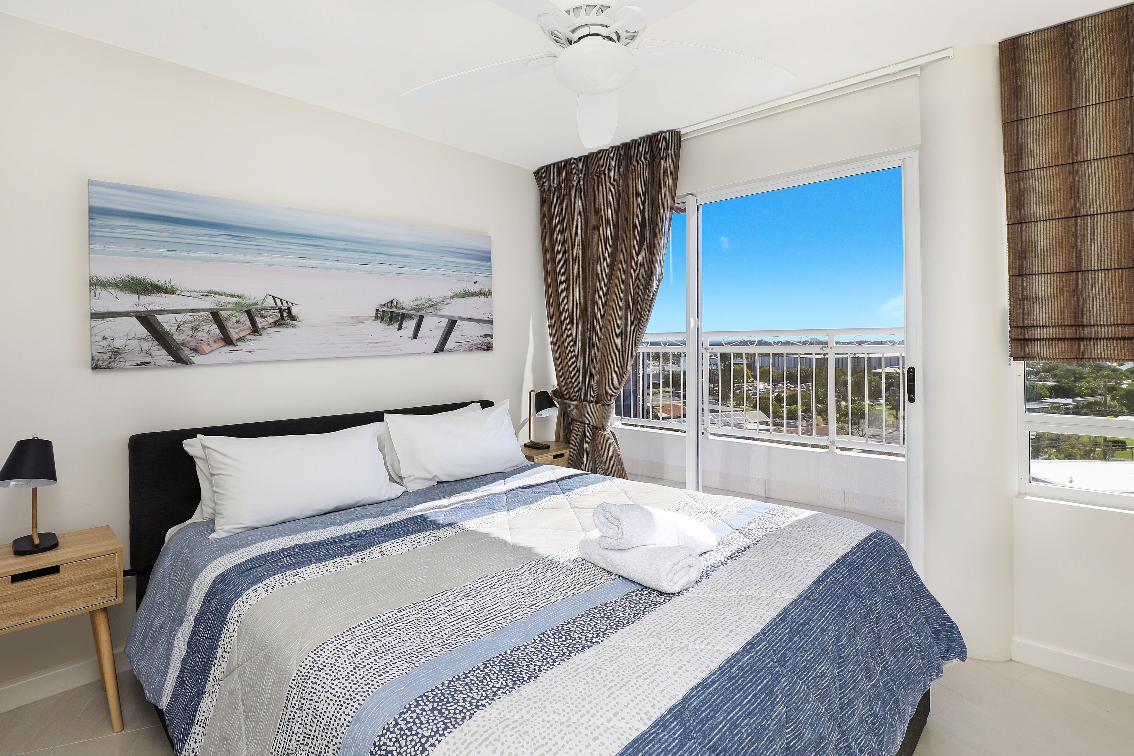 A bedroom with a bed, a nightstand, a window with curtains, and a painting of a beach scene.