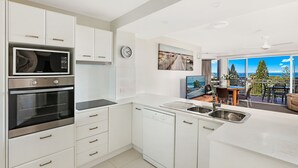 A modern kitchen with white cabinetry, a built-in microwave, oven, and dishwasher. There is a large window offering a view of a balcony with outdoor furniture.