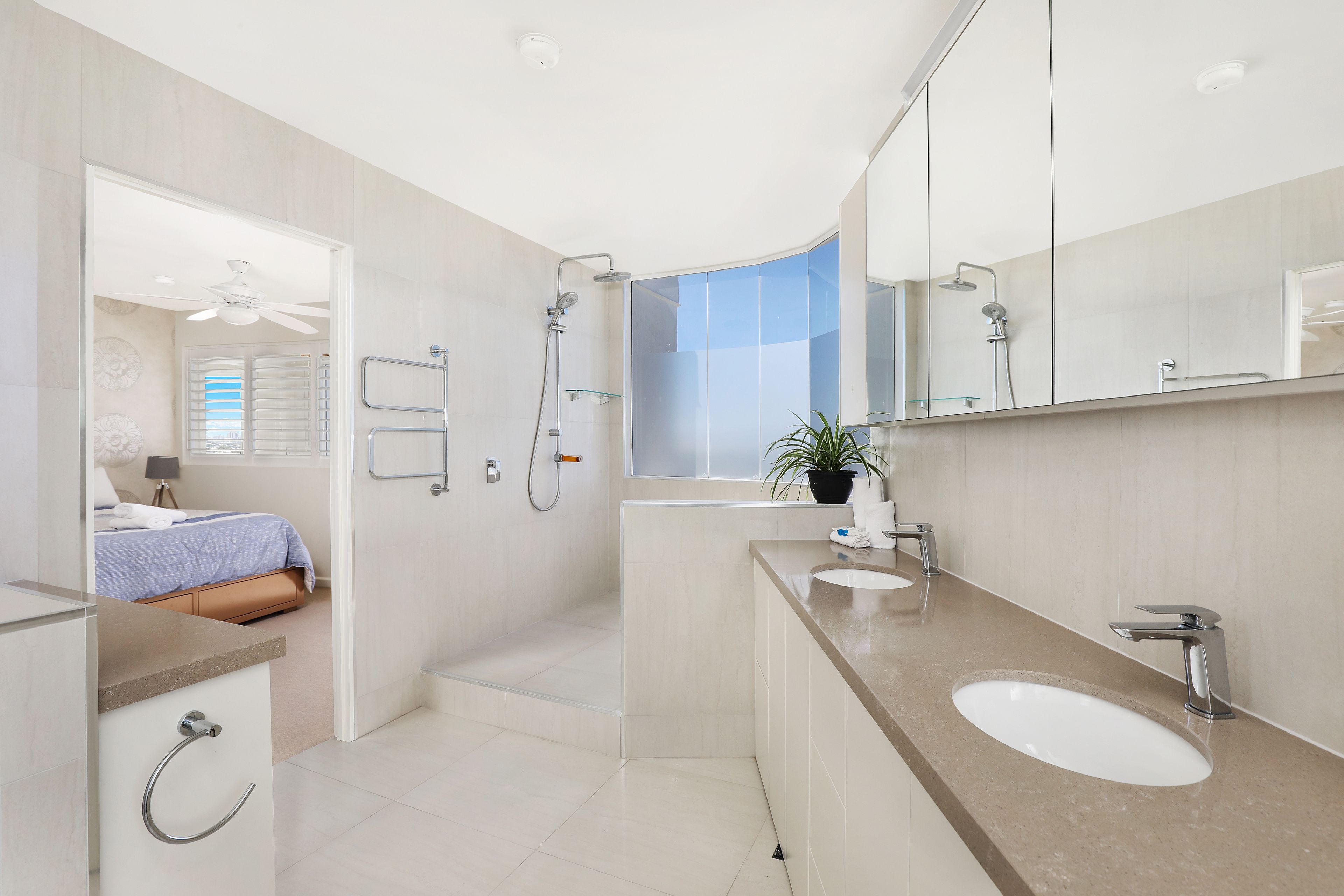 Un baño moderno con un espejo grande, dos lavamanos, una ducha y un ventilador de techo.